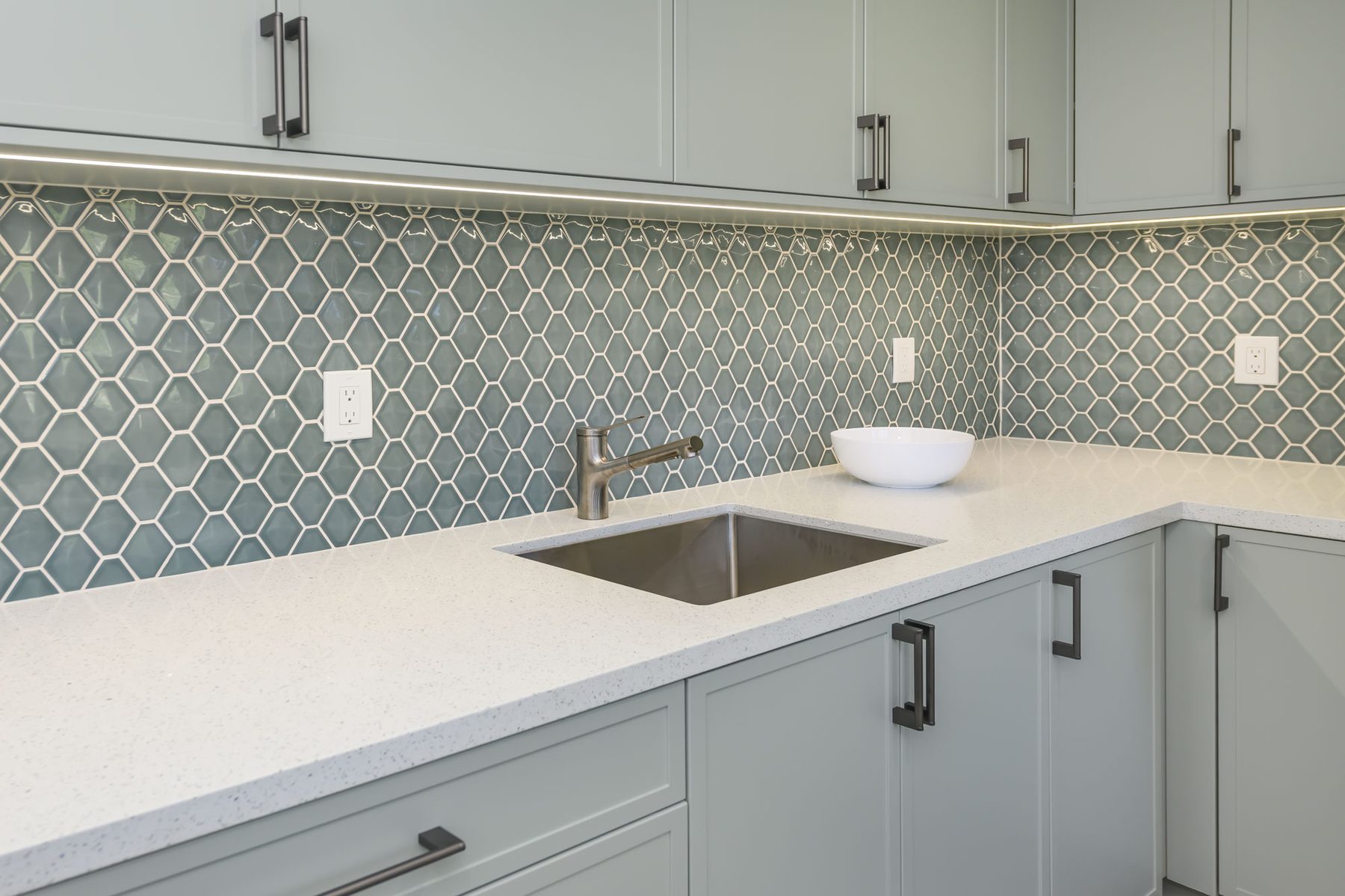 Laundry sink detail with elongated green-blue honeycomb glass mosaic, undermount stainless sink, and quartz counter