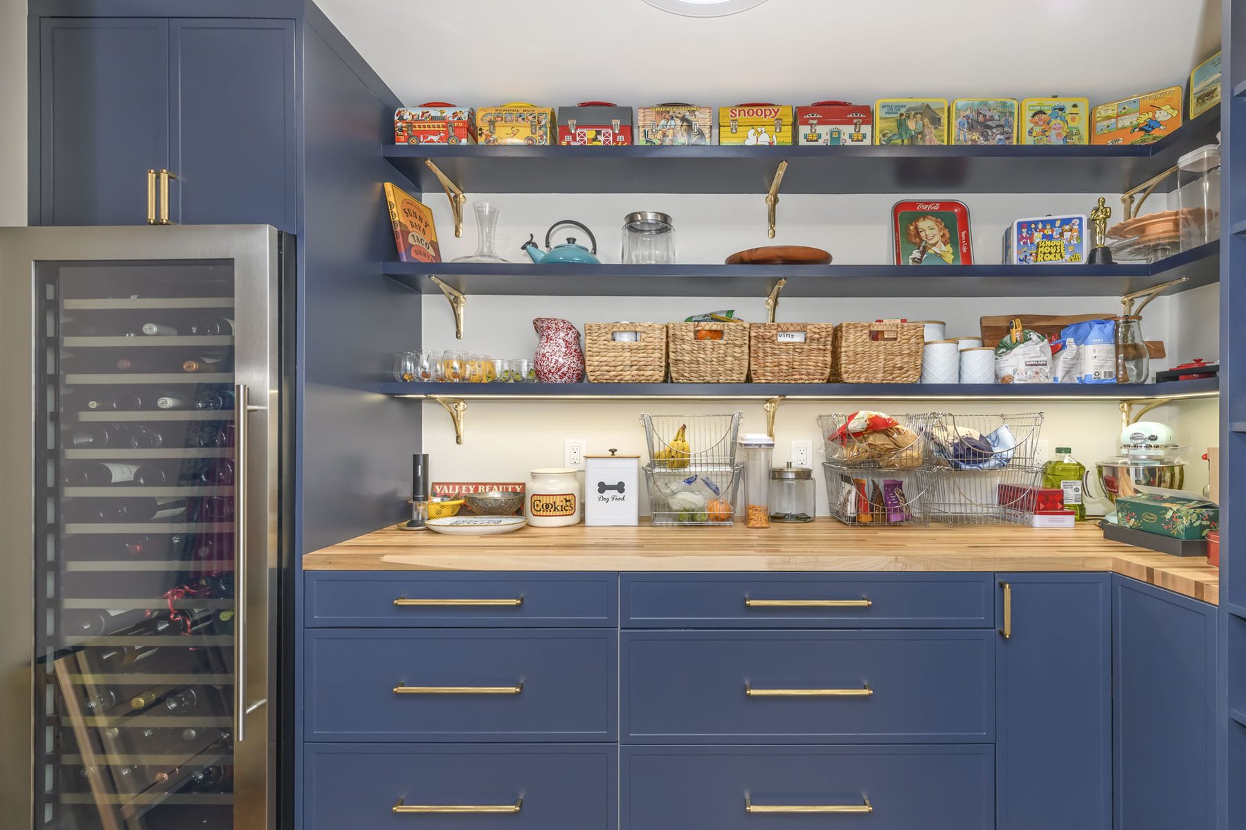 Working pantry with navy cabinetry, butcher-block counter, vintage tin lunch box collection on open shelving, and built-in wine column