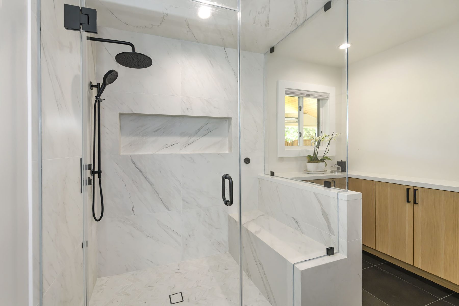 Detail of primary shower with marble niche, herringbone floor mosaic, black rainhead, and integrated marble bench